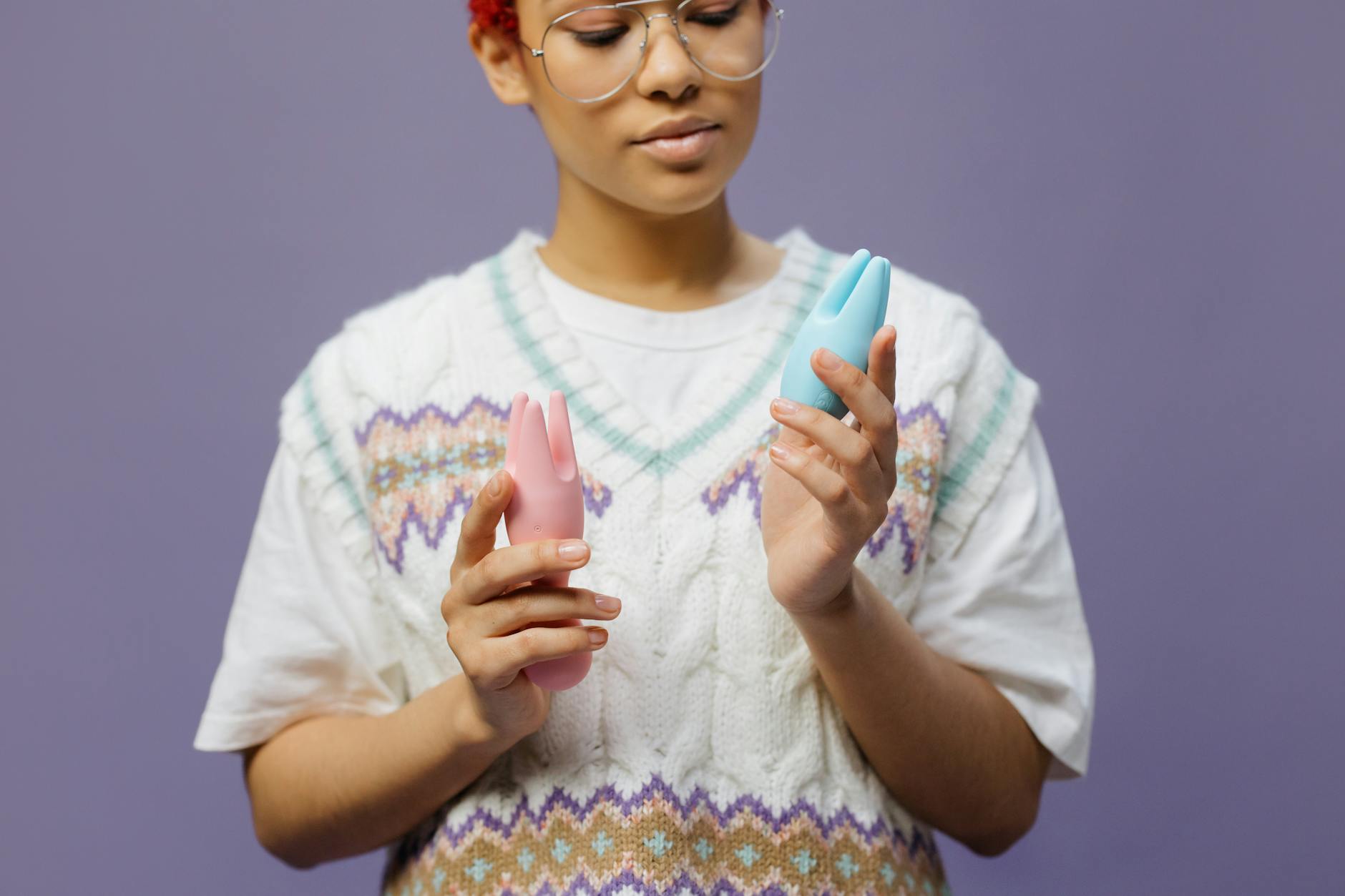 Woman holding blue and pink silicone vibrators in a contemplative pose against neutral background